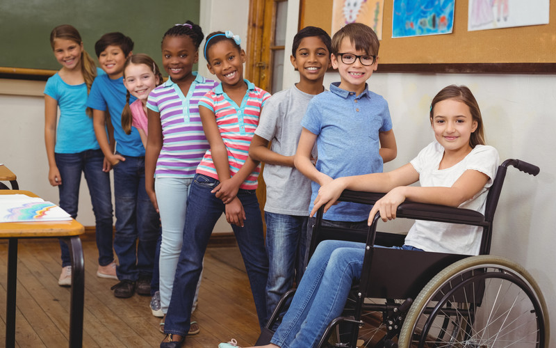 En gruppe elever på barnetrinnet står foran ei tavle i et klasserom. En av elevene sitter i rullestol.