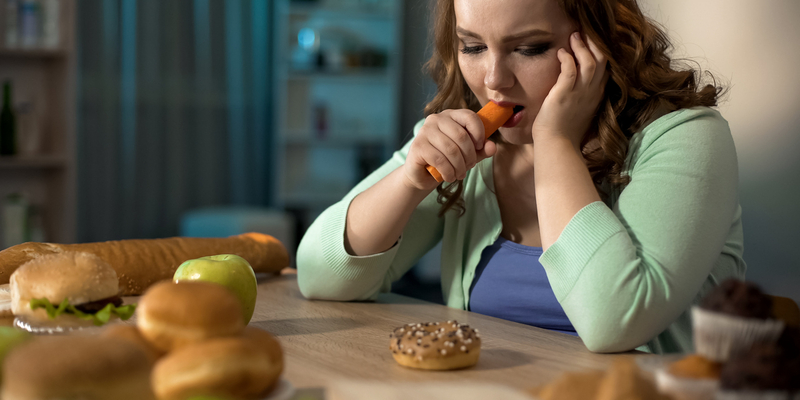 Frustrert kvinne sitter ved spisebord og spiser på en rå gulrot, men det ligger bakverk og annet mindre sunn mat på bordet foran henne.