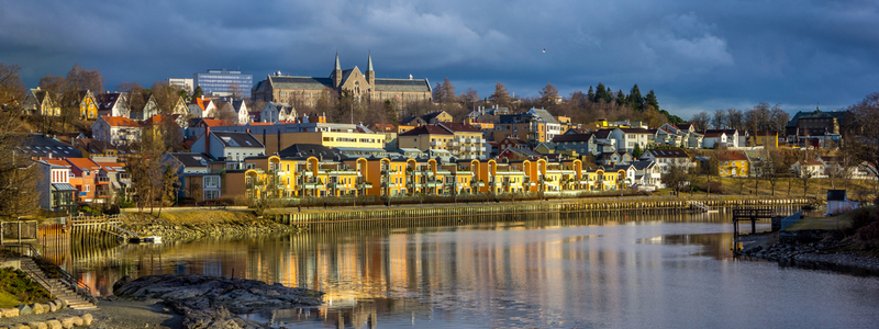 Bilde fra Trondheim hvor man ser bl.a. Nidelva i forgrunnen, forskjellige typer bygninger - alt fra eneboliger, til boligkomplekser, næringsbygg og øverst ser man flere NTNU-bygg på Gløshaugen.