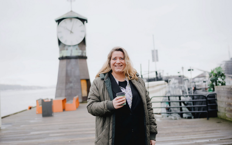 Smilende kvinne med kaffekopp på en brygge med sjø og et klokketårn bak.