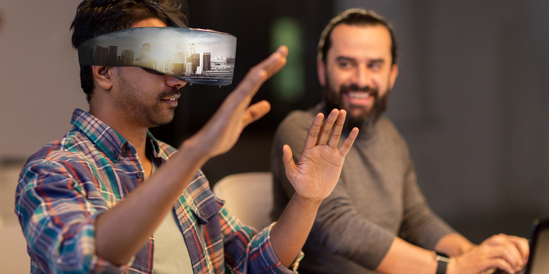 To menn sitter foran en PC, den ene bruker en VR-brille og holder hendene opp foran seg.