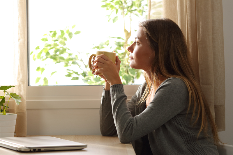 Ung kvinne som ser tankefullt ut vinduet med et kaffekrus i hånda.