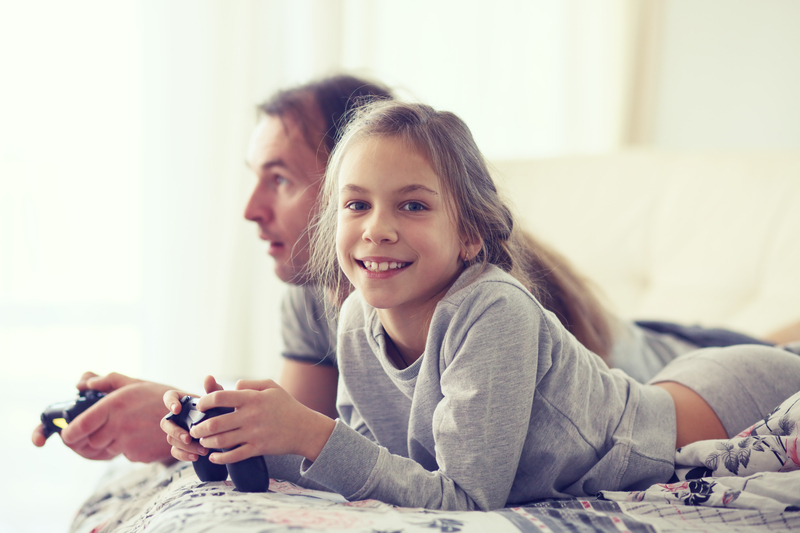 Far og datter som spiller TV-spill med hver sin konsoll i hånda. Datter smiler rett inn i kamera.